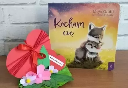 Zdjęcie przedstawiajace książeczkę pod tytułem Kocham Cię oraz pracę plastyczną w postaci papierowych, czerwonych serc
